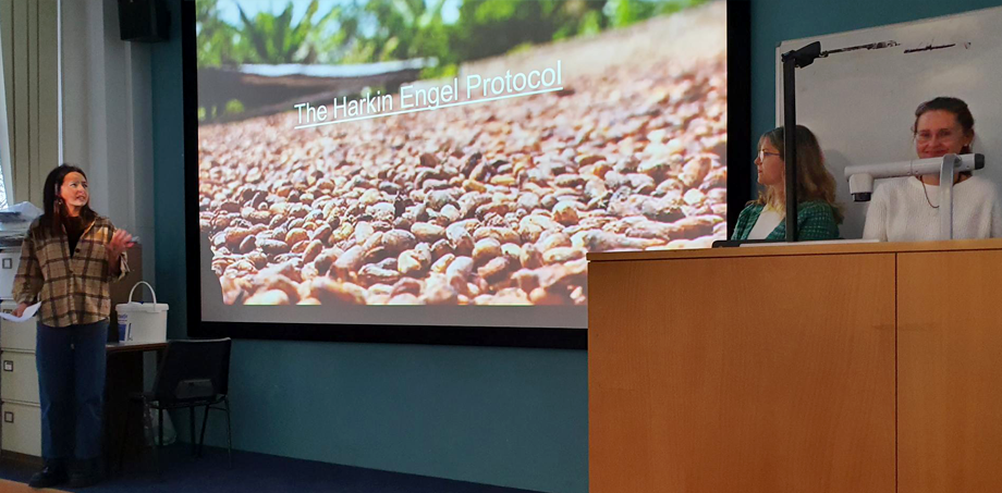 Photo: A female student of the group talking to the classroom with a slide projected behind her showing cocoa beans drying in the sun, entitled: The Harkin Engel Protocol.