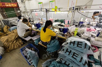 Photo: Young people using sewing machines at work stations aligned next to one another on a long table surrounded by bags of material in a garment factory that appears to be asian.