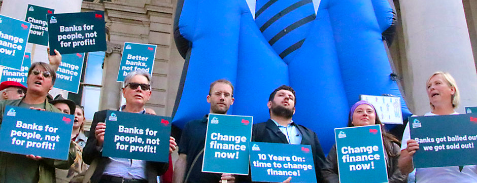 Photo: Adults demonstrating in public in front of a building with an inflated blue suit. They are holding printed cards with the text: Banks for people, not for profit; Change finance now; 10 ears on: time to change finance, and Banks fot bailed out we got sold out!.
