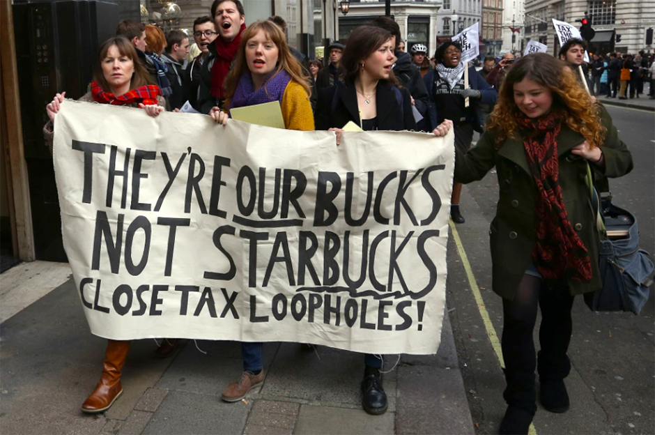 Photo: Adult women demonstrating on the streets leading a group of protestors, holding a banner with the text: They're our bucks not Starbucks, close tax loopholes!