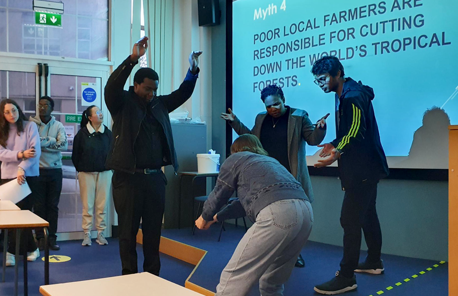 Photo of members of the group presenting a dramatisation of Myth 4, poor famers responsible of cutting down the forest. Female member pretends to use an axe to cuts down a tree represented by another member standing with arms over his head. Others watch and ask why.